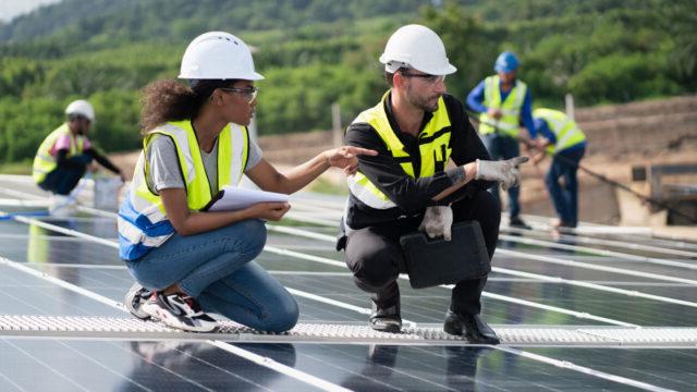 Two people wearing yellow vests and hard hats crouching on solar panels