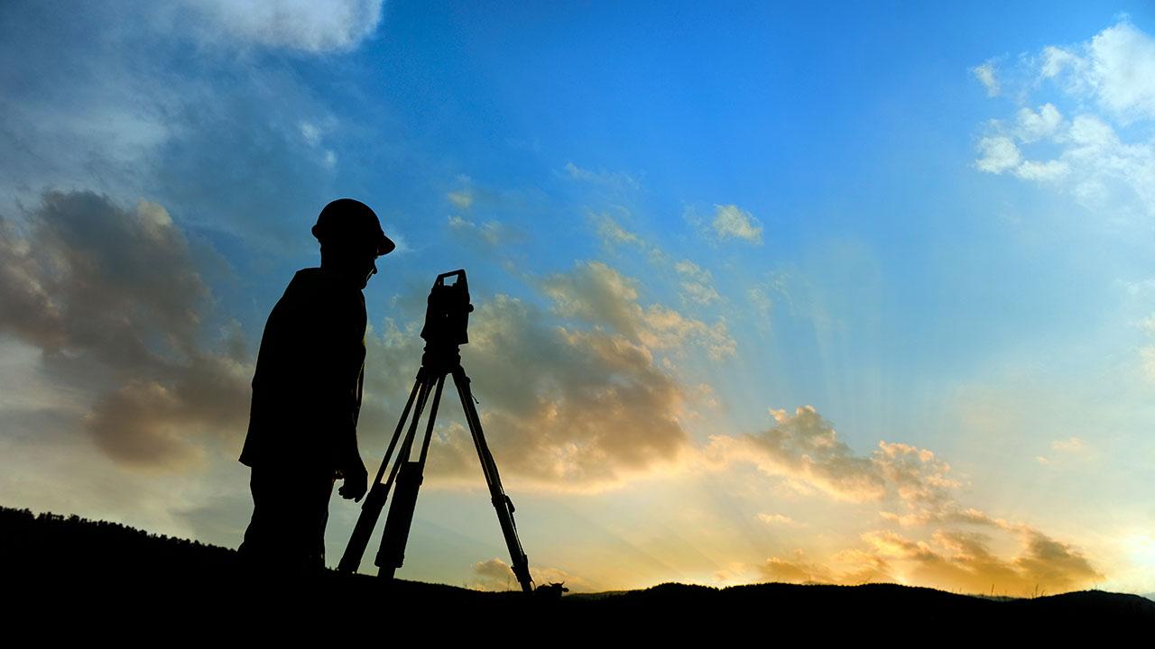 Silhouette of a person wearing a hard hat looking through a total station