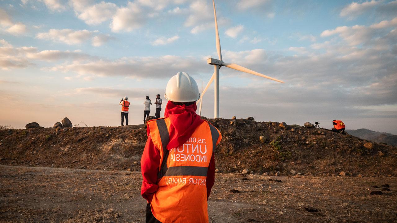 A female wind power engineer stands looking at a windmill in the distance in Indonesia