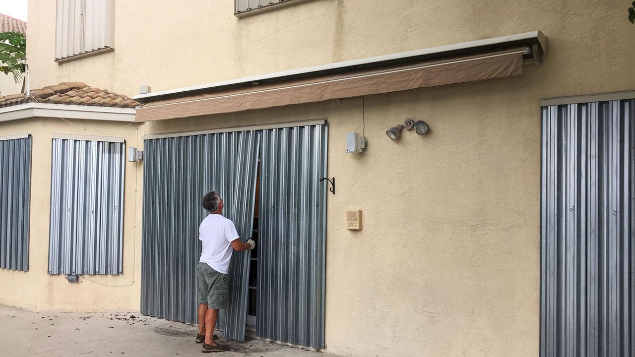 Man wearing gloves installing metal hurricane shutters on tan building