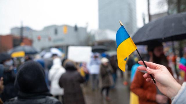 A hand flying a small flag of Ukraine against a blurred urban backdrop